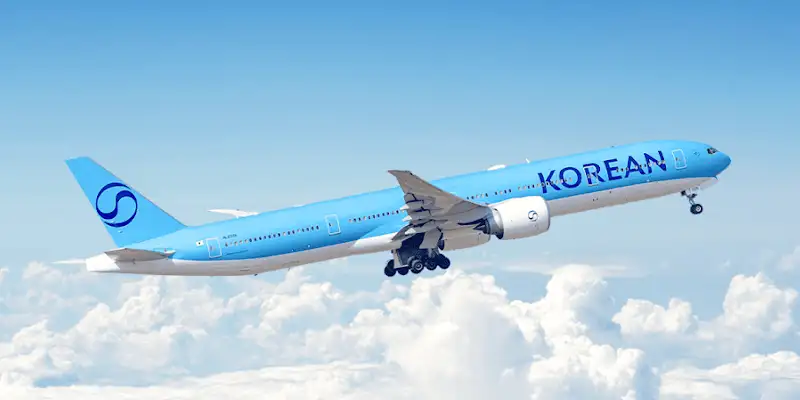 Korean Air Boeing 777 aircraft ascending with landing gear retracted against a backdrop of clouds and blue sky.